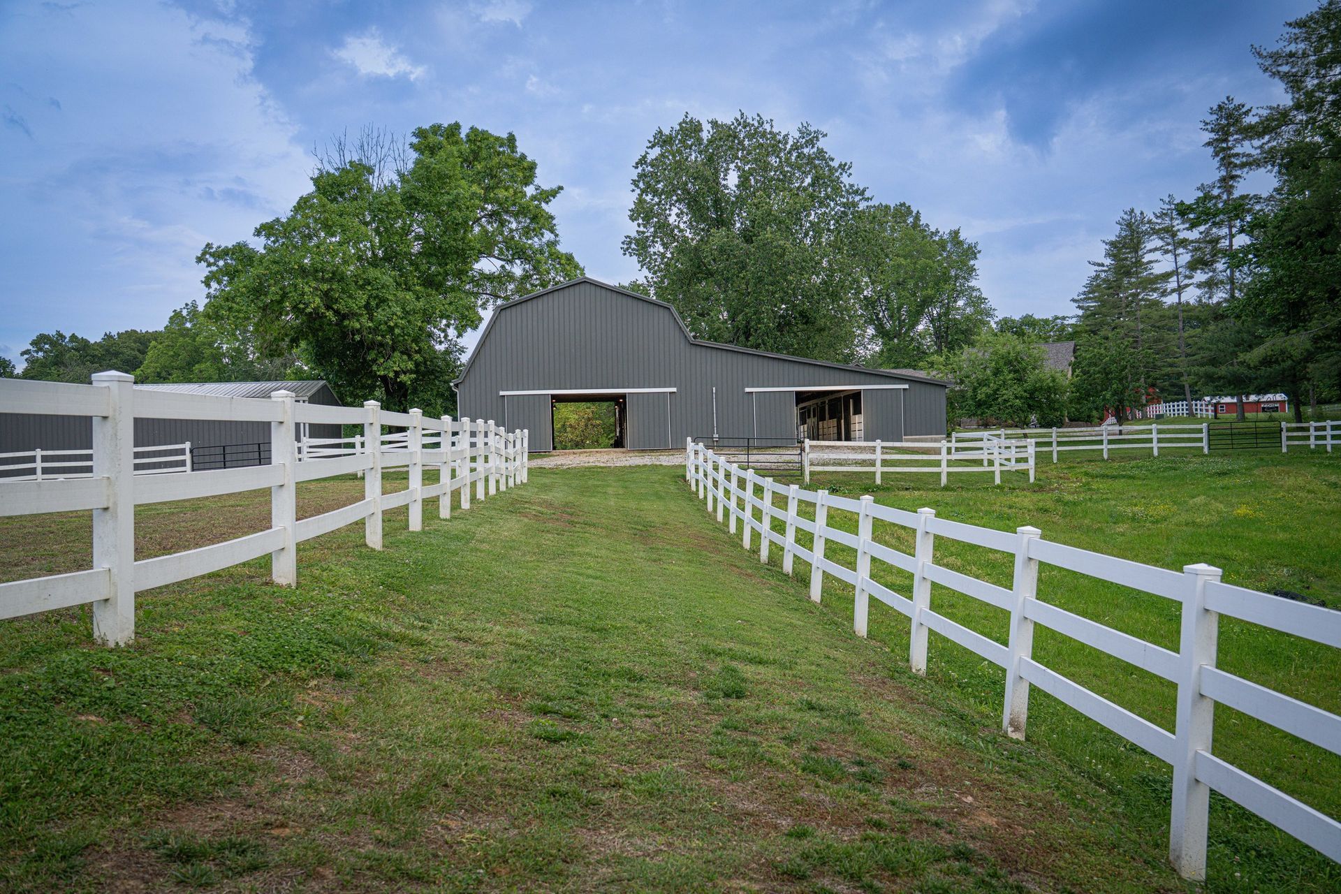 Gray barn with open doors, flanked by white fences in a grassy field with trees under a blue sky.