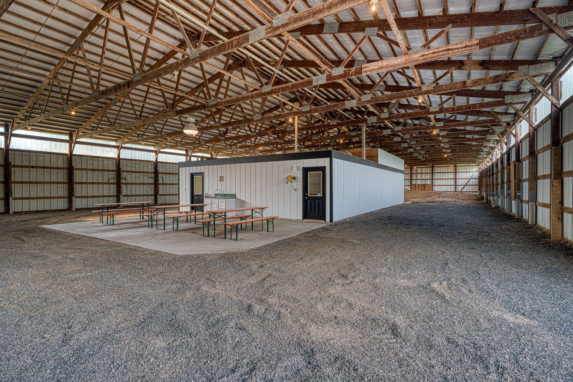 Interior of an open-sided barn with a gravel floor. A white building with a black door and picnic tables are in the center.
