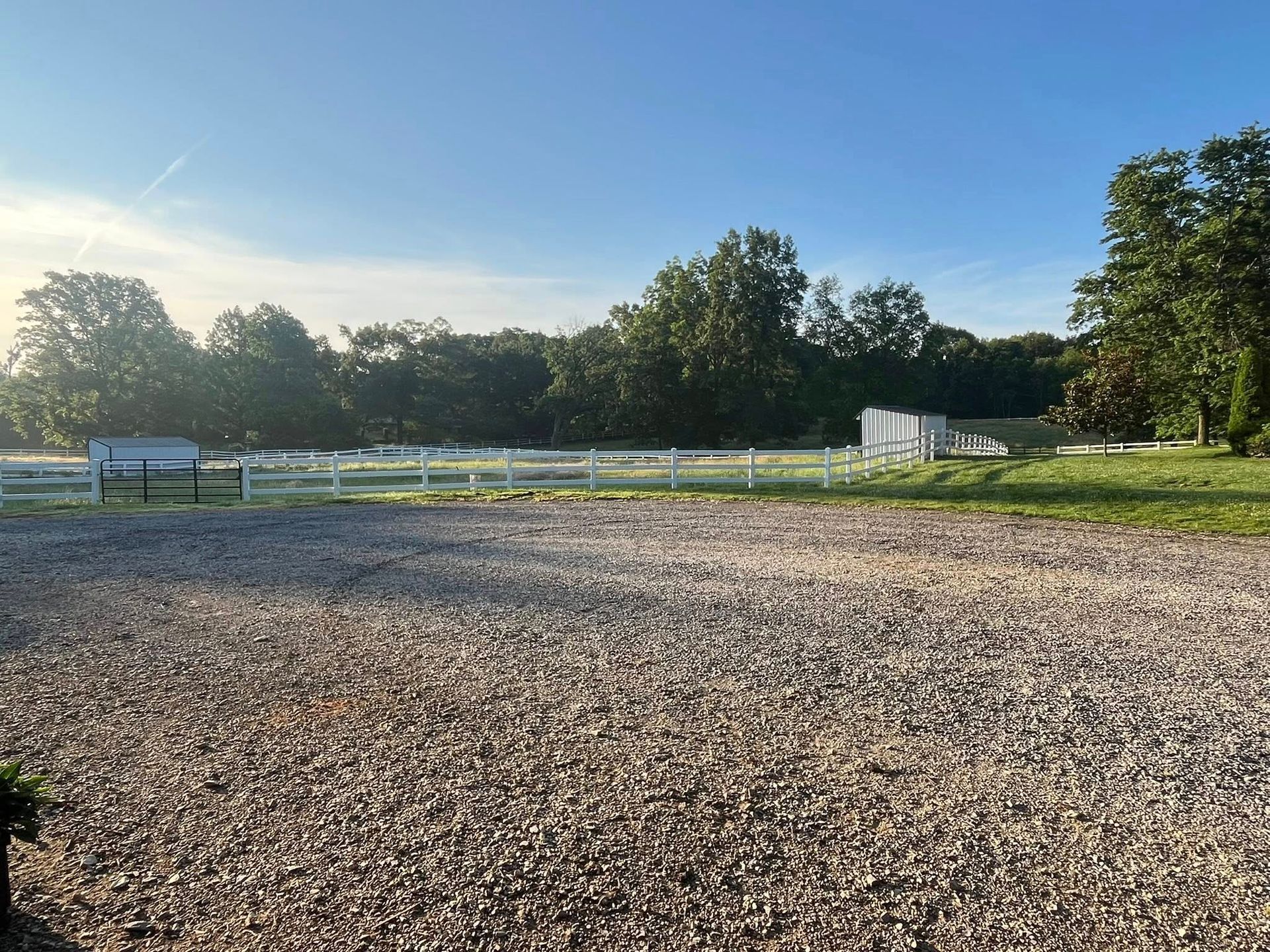 A gravel area in front of a grassy field with a fence, trees, and a bright blue sky.