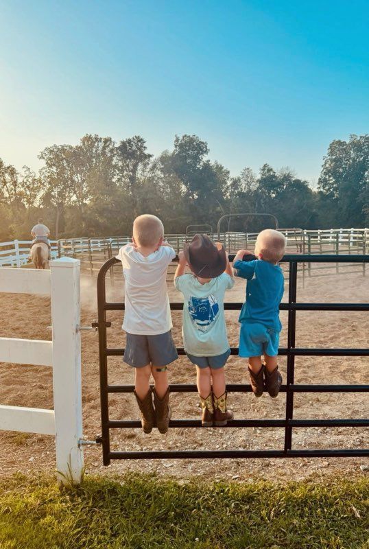 Three young children in cowboy boots perched on a fence, looking at a corral.