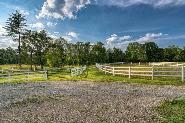 White-fenced horse corral on grassy land, gravel driveway, trees, blue sky, clouds.