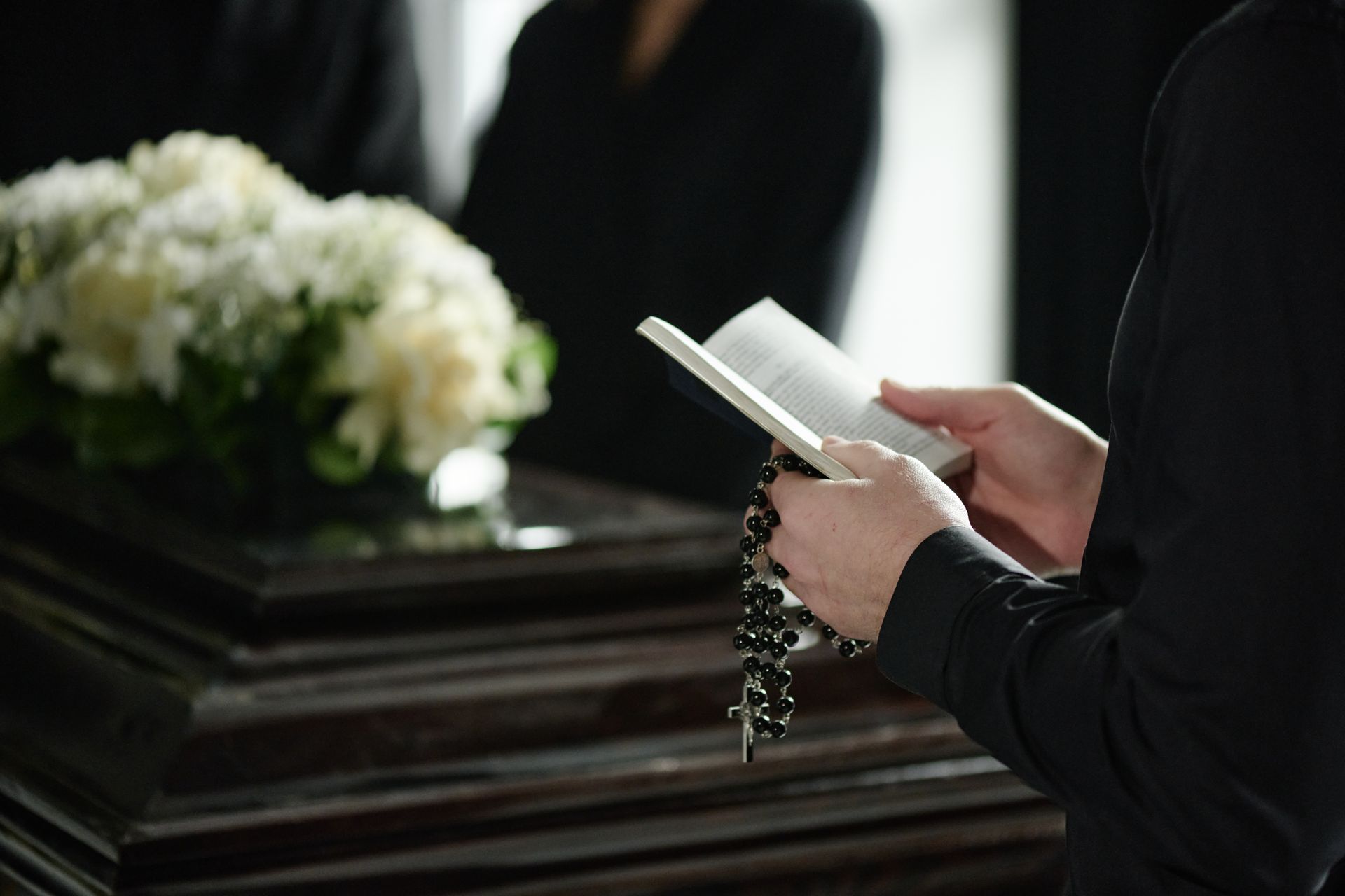 Person holding a prayer book and rosary beside a casket with white flowers