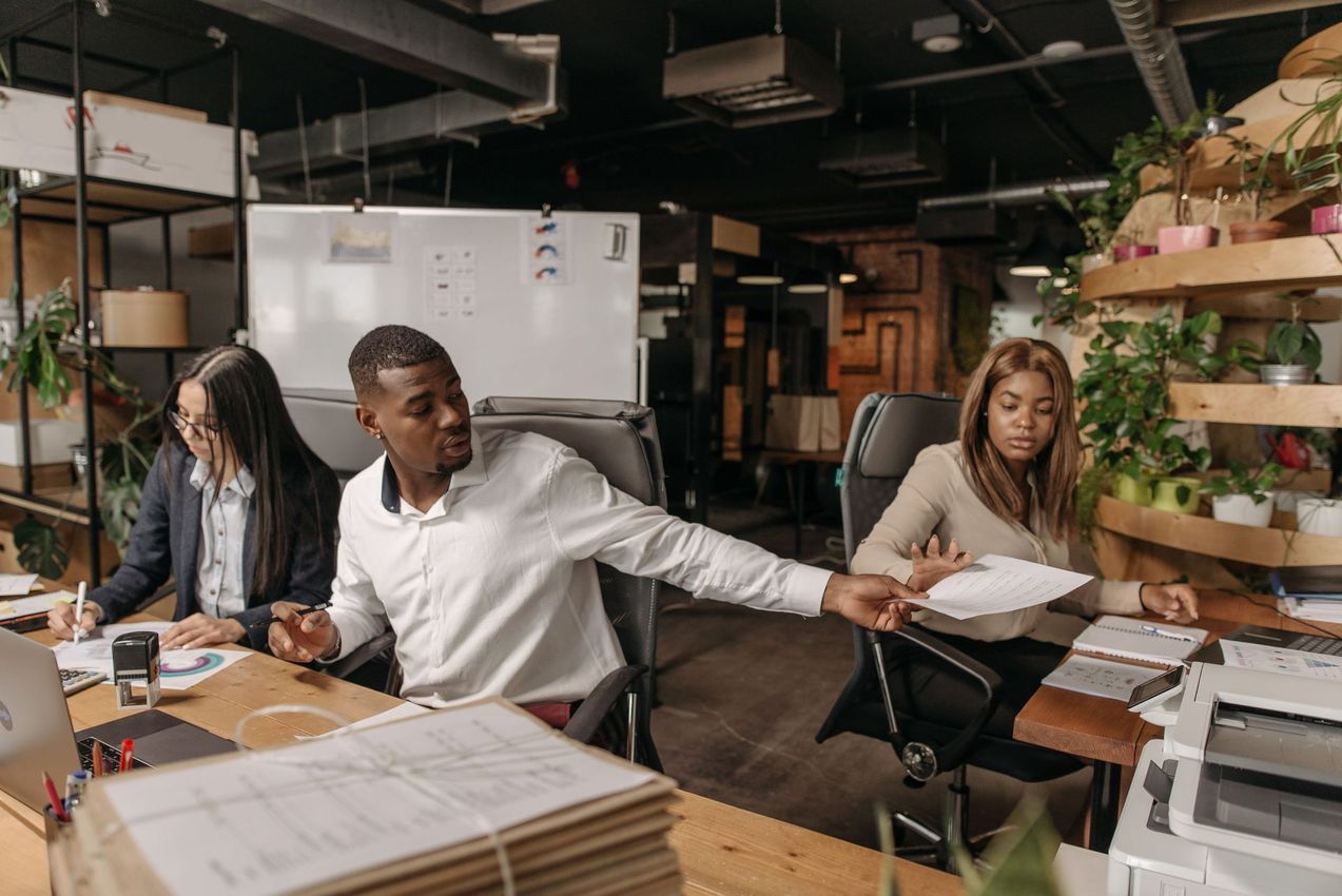 Three coworkers in an office gather around a desk with papers and a printer in an open workspace.