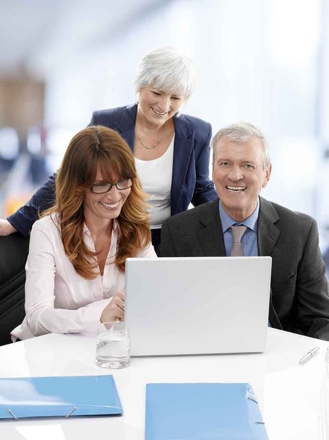 Three coworkers smiling and using a laptop at a bright office table with blue folders.