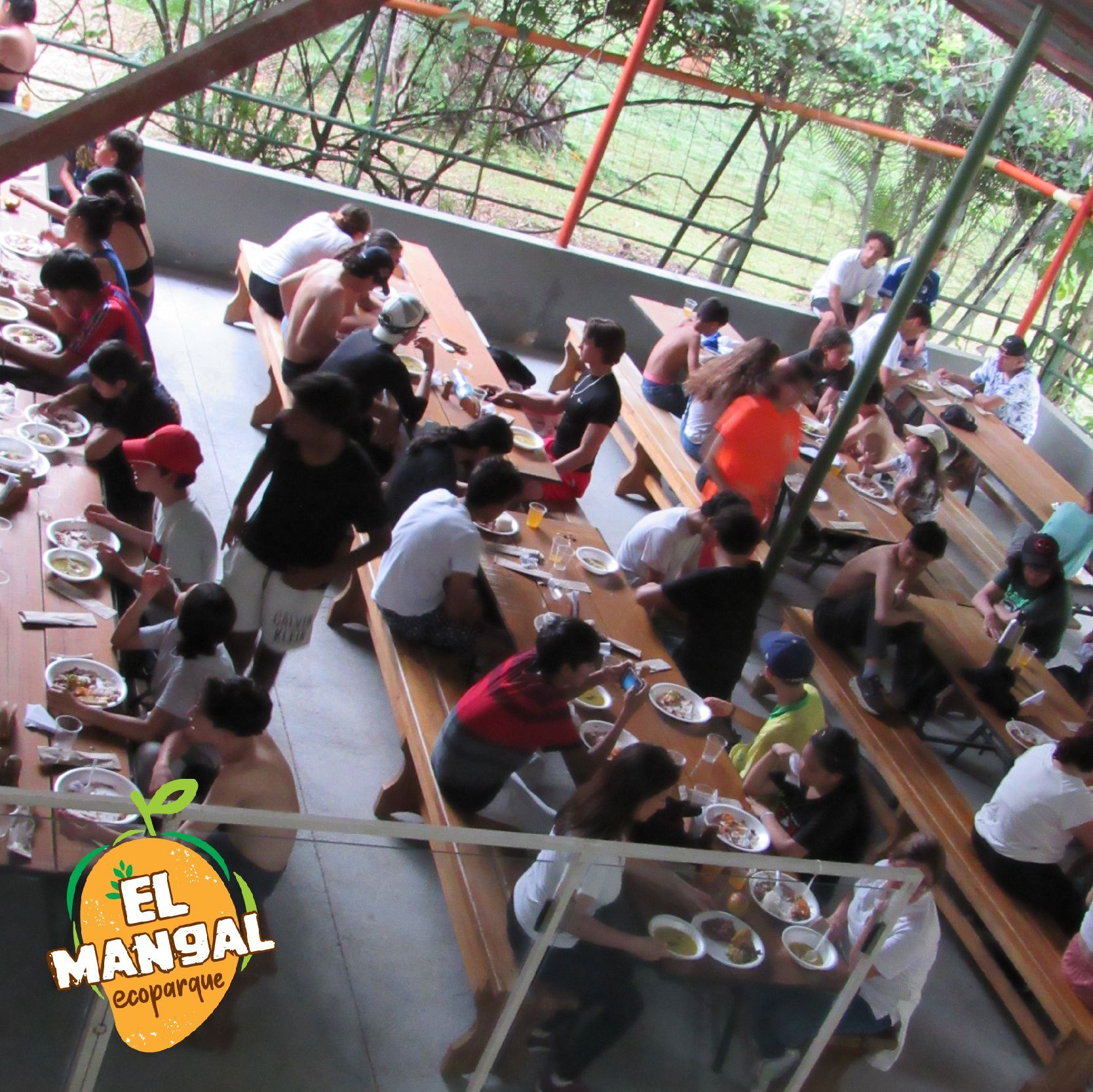 Personas comiendo en largas mesas de madera al aire libre. El logo de 