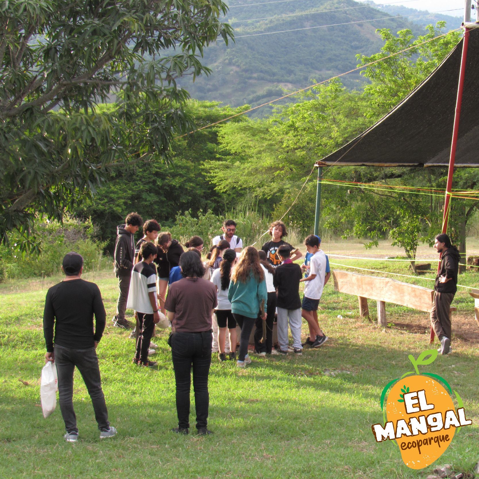 Grupo de personas reunidas al aire libre bajo una estructura de dosel cerca de una montaña.