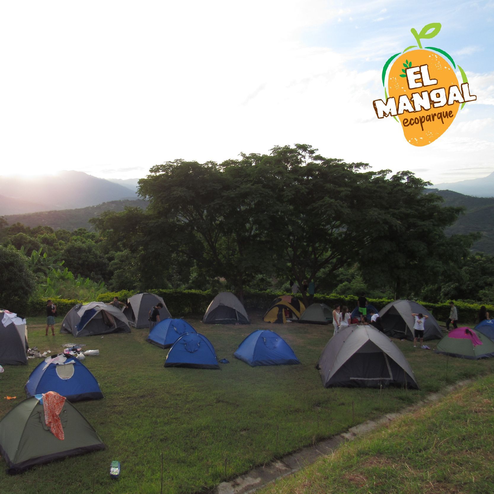 Camping con tiendas de campaña en una colina con césped, con las montañas al fondo. El logo de 