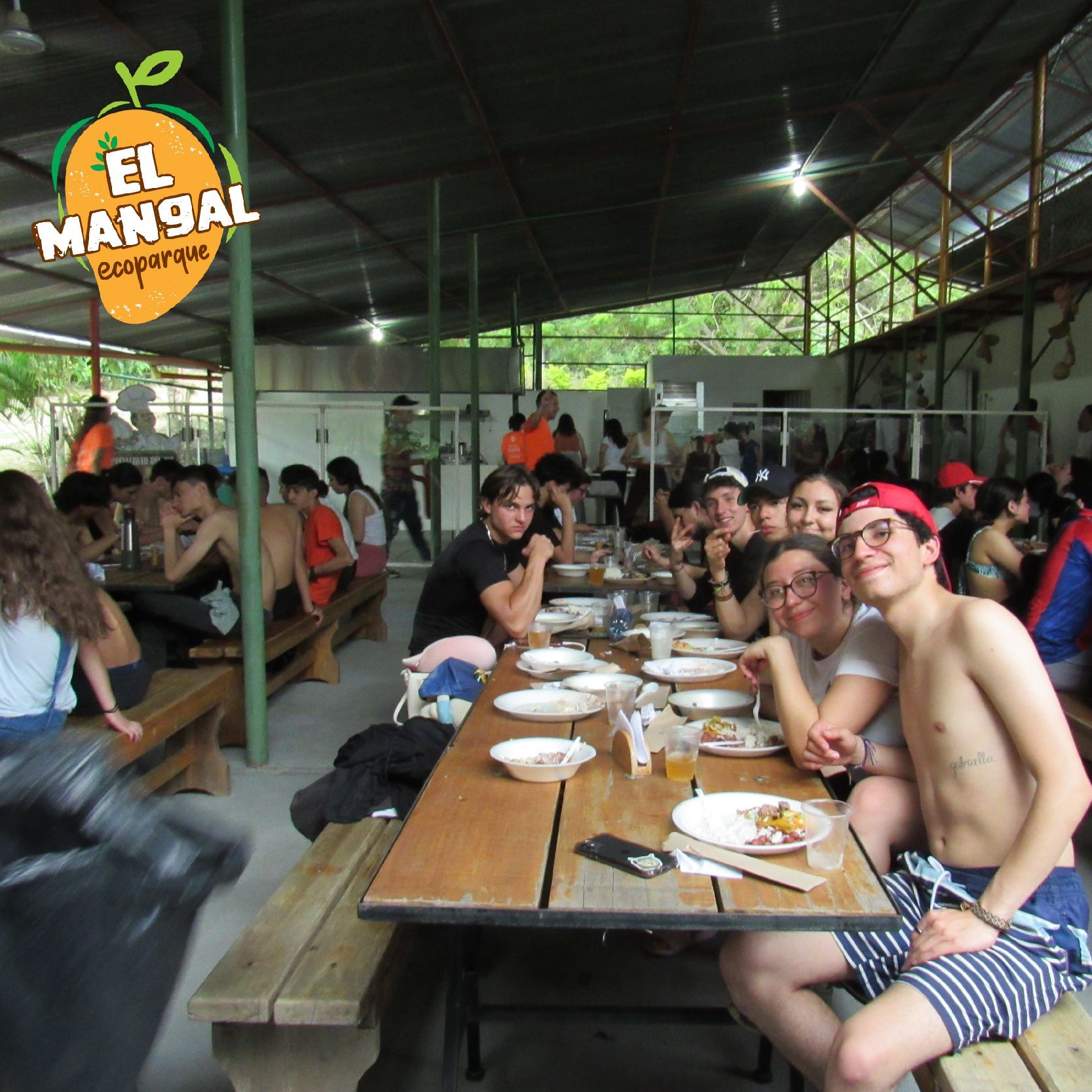 Personas cenando en el Ecoparque “El Mangal”, sentadas en mesas de madera, comiendo bajo una estructura techada.