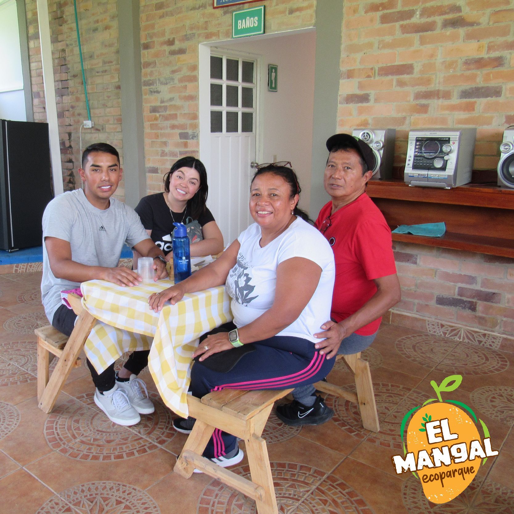 Cuatro personas sentadas en una mesa de picnic. Exterior con pared de ladrillo, radio y puerta abierta.