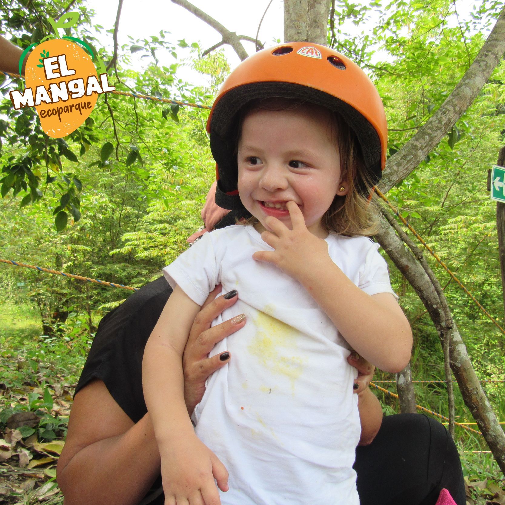 Chica con casco naranja y camisa sucia, sujeta por una persona. Fondo de bosque.