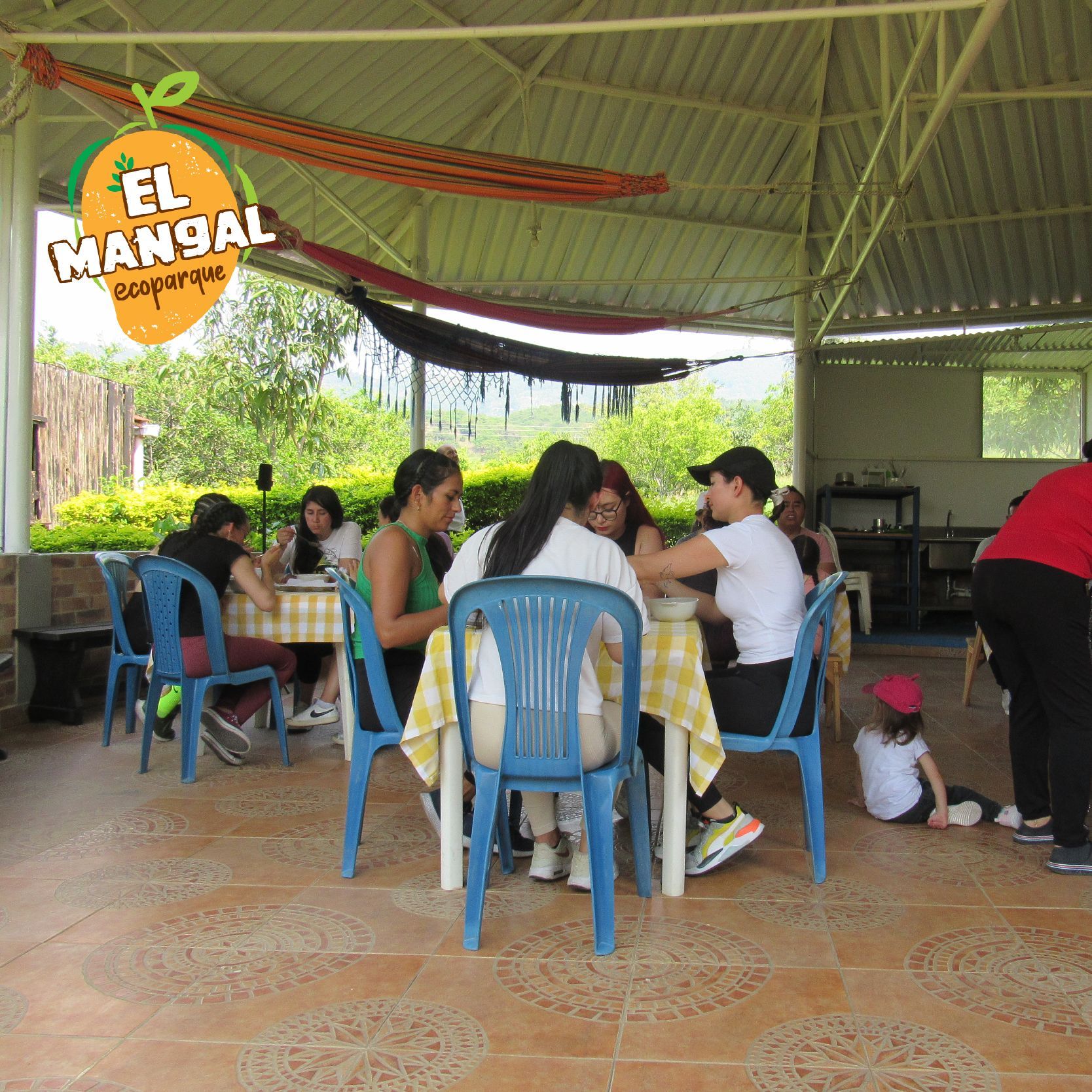 Personas sentadas a la mesa comiendo en una zona exterior cubierta con hamacas. El área tiene el logo de 