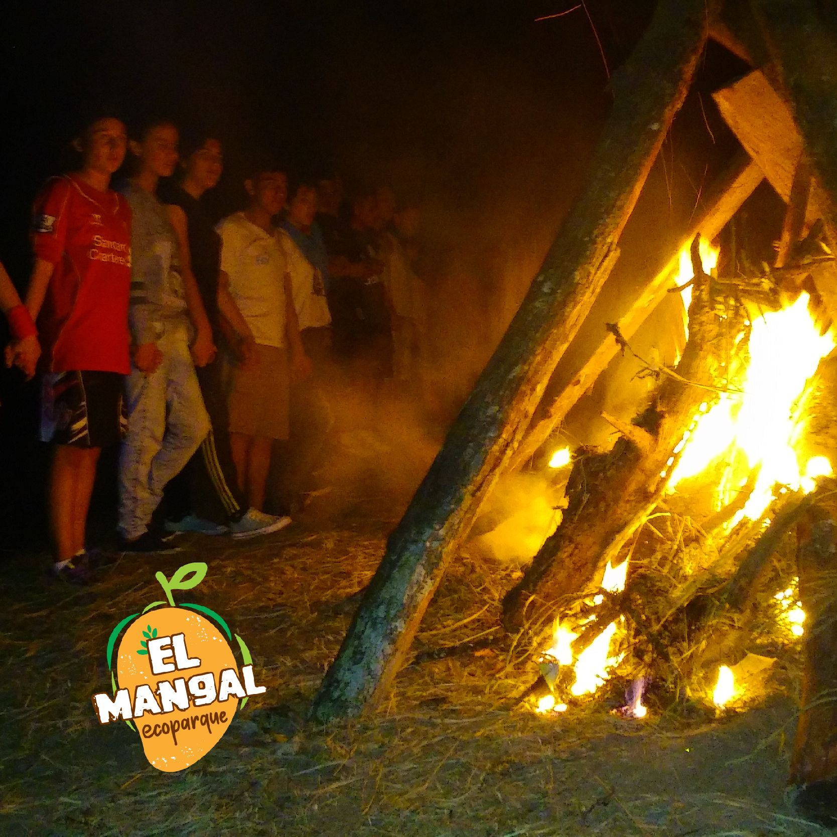 Gente de pie cerca de una hoguera por la noche; llamas anaranjadas iluminan la escena.