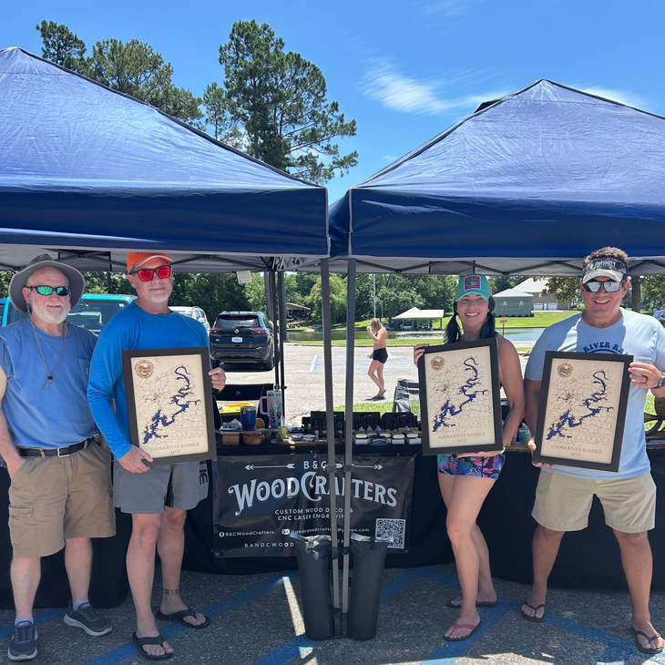 Four people under blue canopies at an outdoor market, holding framed art prints and standing by a sign reading “WoodCrafters.”