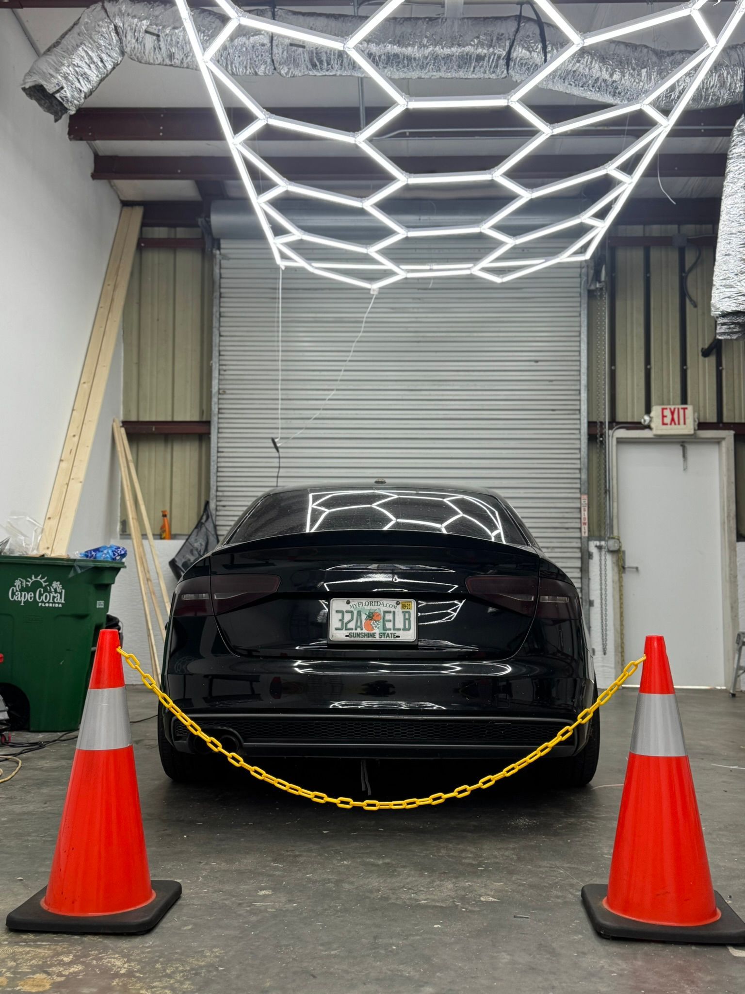 Black car behind orange cones and chain barrier in a garage-like bay with overhead lights