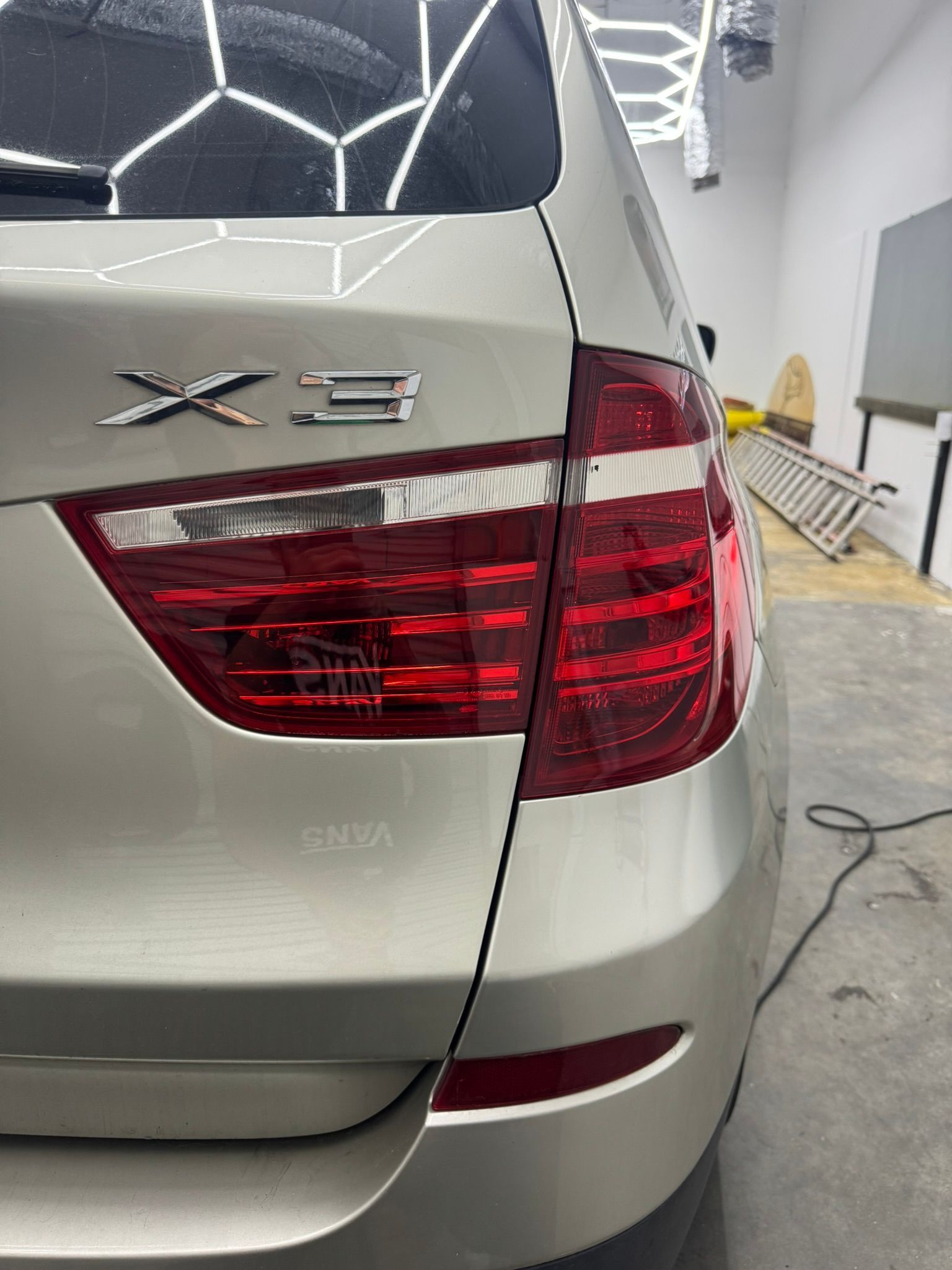 Rear left of a silver BMW X3, showing the taillight in a garage or workshop.