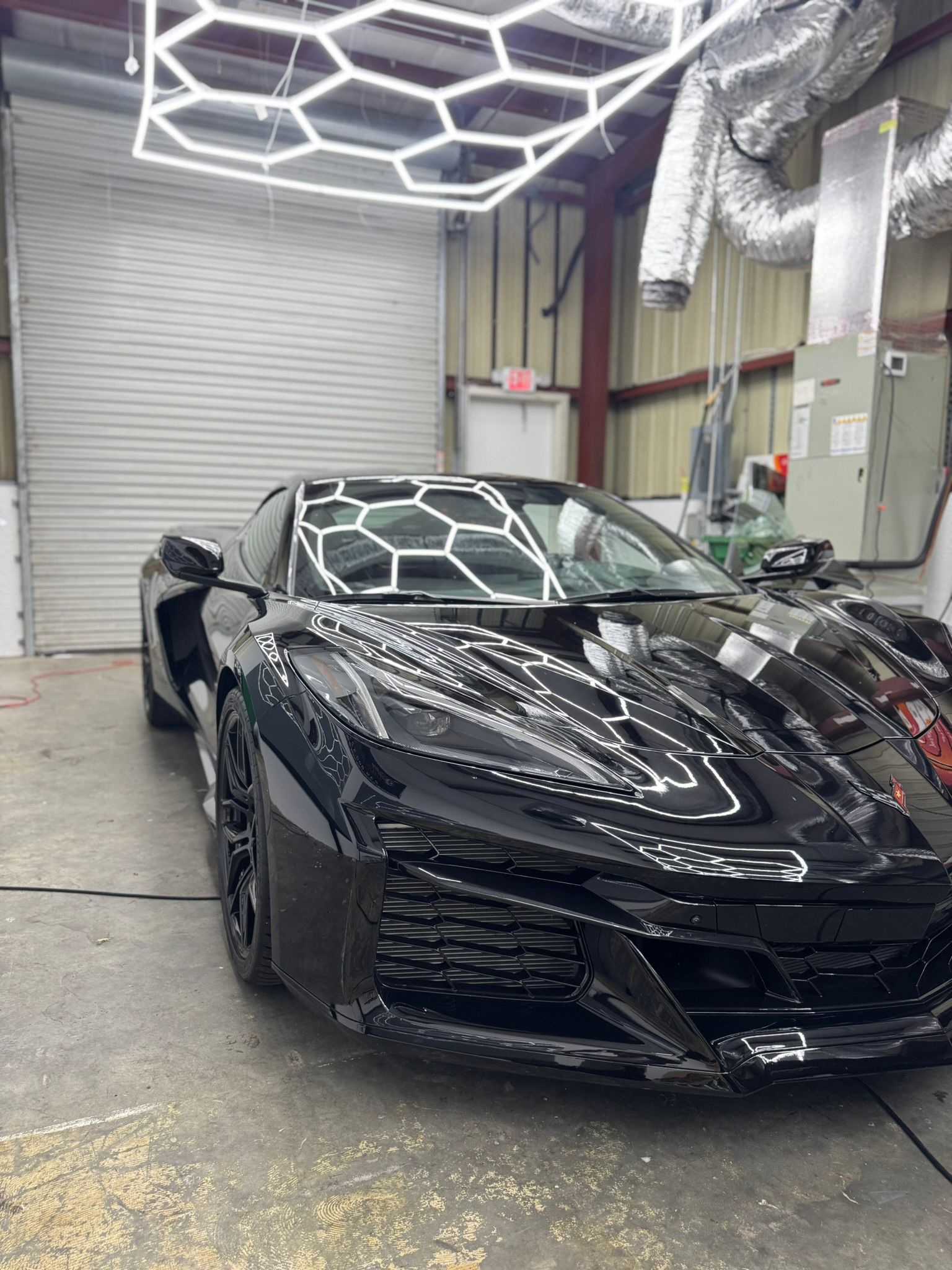 Black sports car in an auto detailing bay with bright geometric ceiling lights reflecting on its hood.