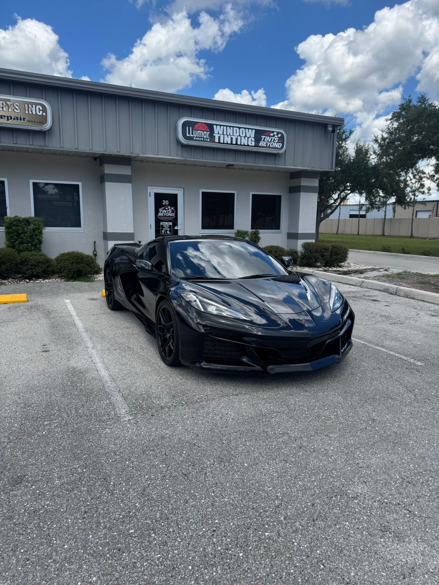 Black sports car parked in front of a gray storefront under a partly cloudy sky.