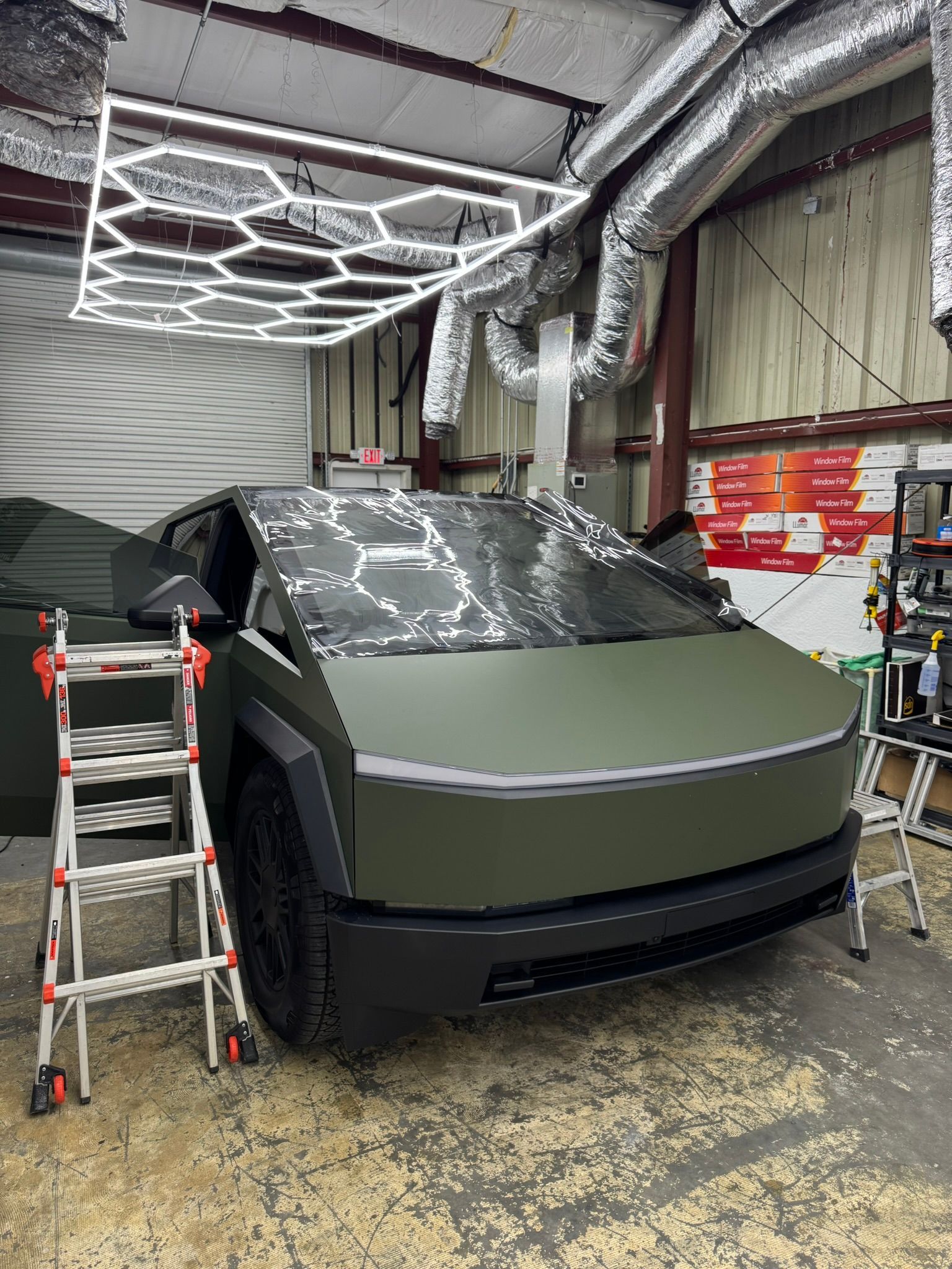 Angular gray Cybertruck indoors beside a ladder, with a light-patterned canopy overhead.