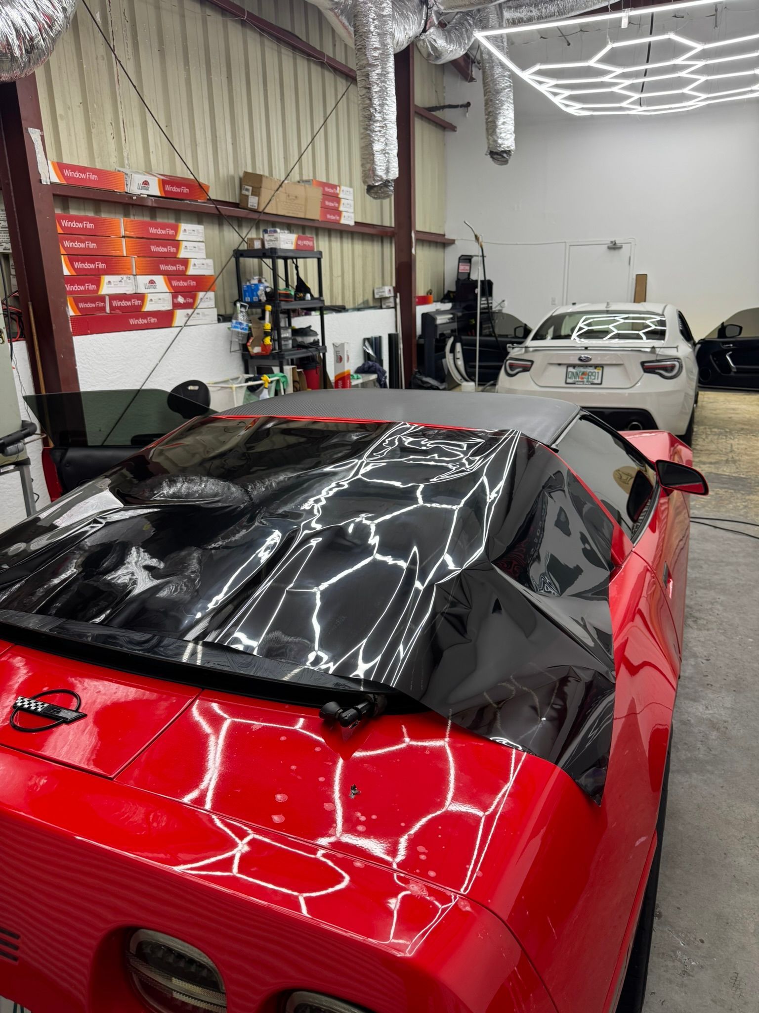 Red sports car with black cover in an auto shop, with a white race car in the background