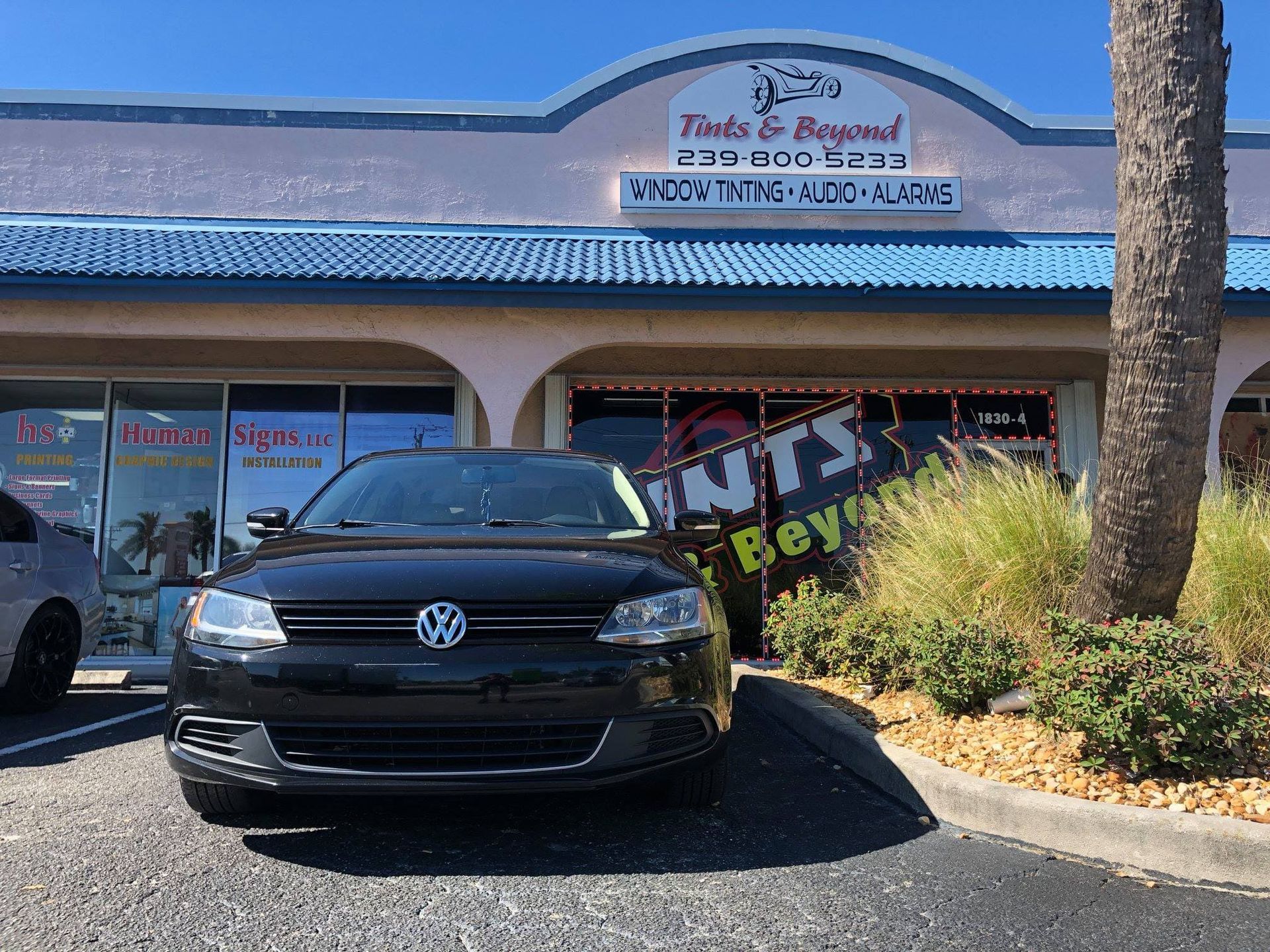 Black Volkswagen parked in front of a storefront with blue awning and palm tree.
