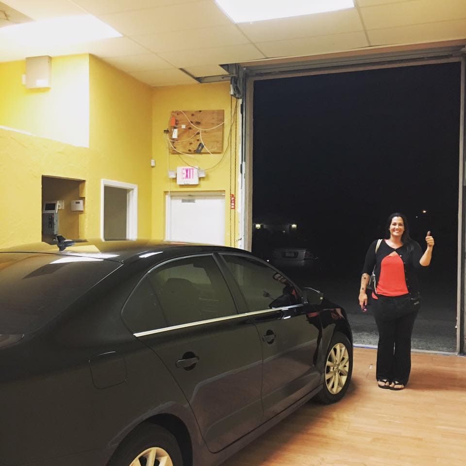Woman standing beside a black car in a yellow-lit garage, smiling and giving a thumbs-up at night