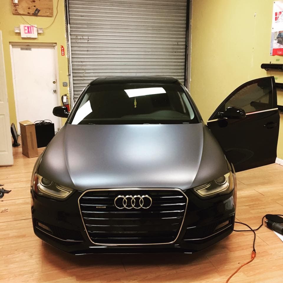 Front view of a matte black Audi in a garage, with the driver door open and a charging cable on the floor