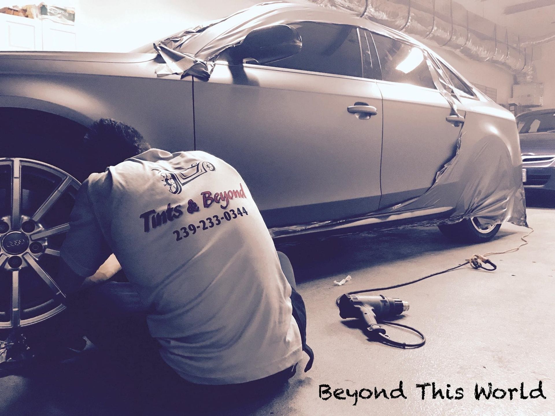 Mechanic sanding a gray car in a workshop, with tools on the floor and “Beyond This World” text.