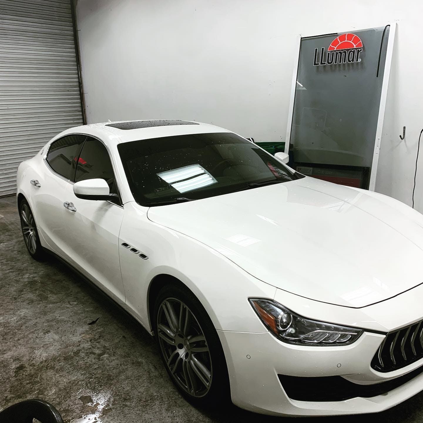 White sedan parked indoors in a garage, angled front-left view with tinted windows and wet concrete floor