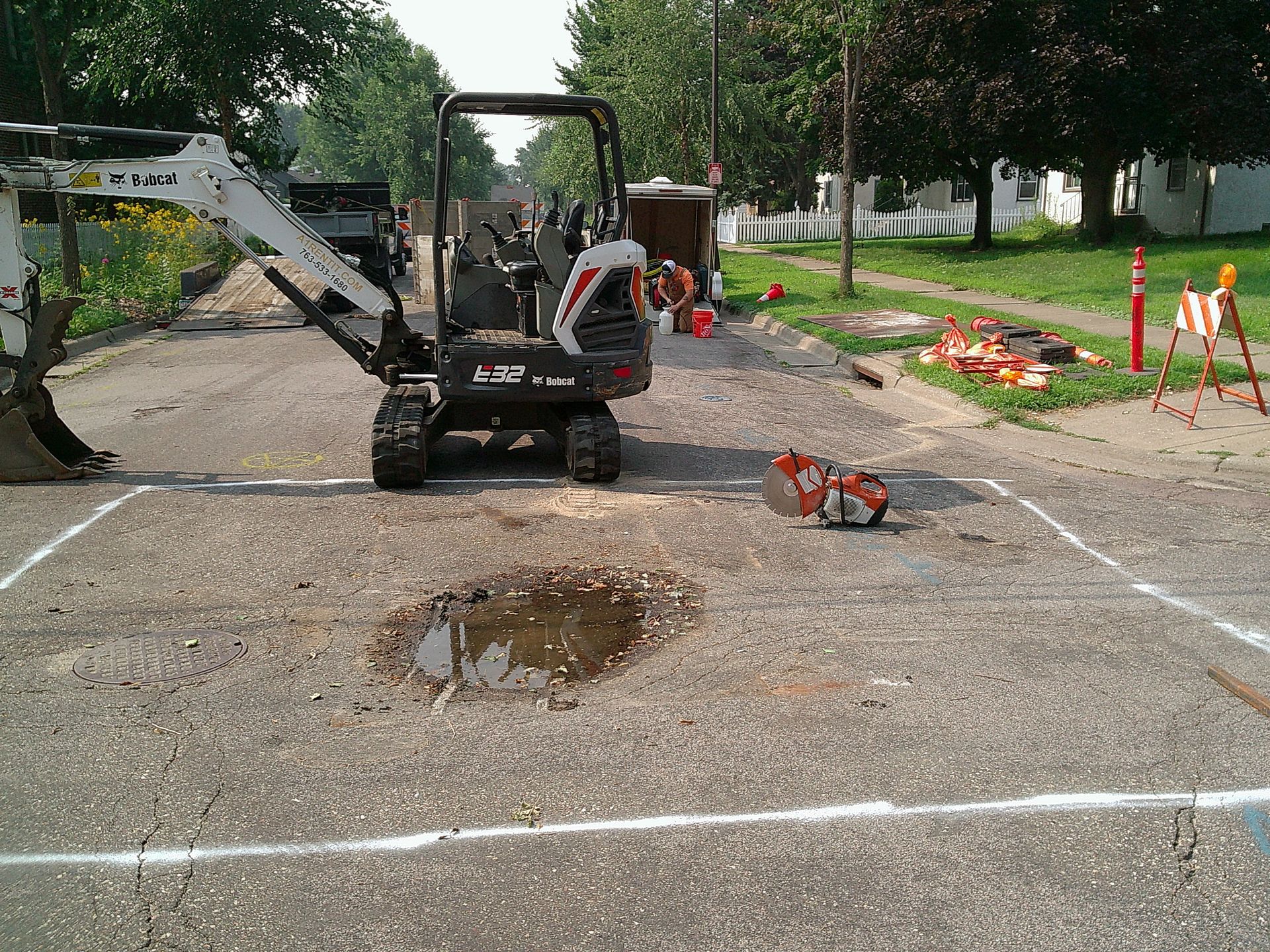 Mini excavator on street construction site, marked area with water puddle, orange traffic cones.