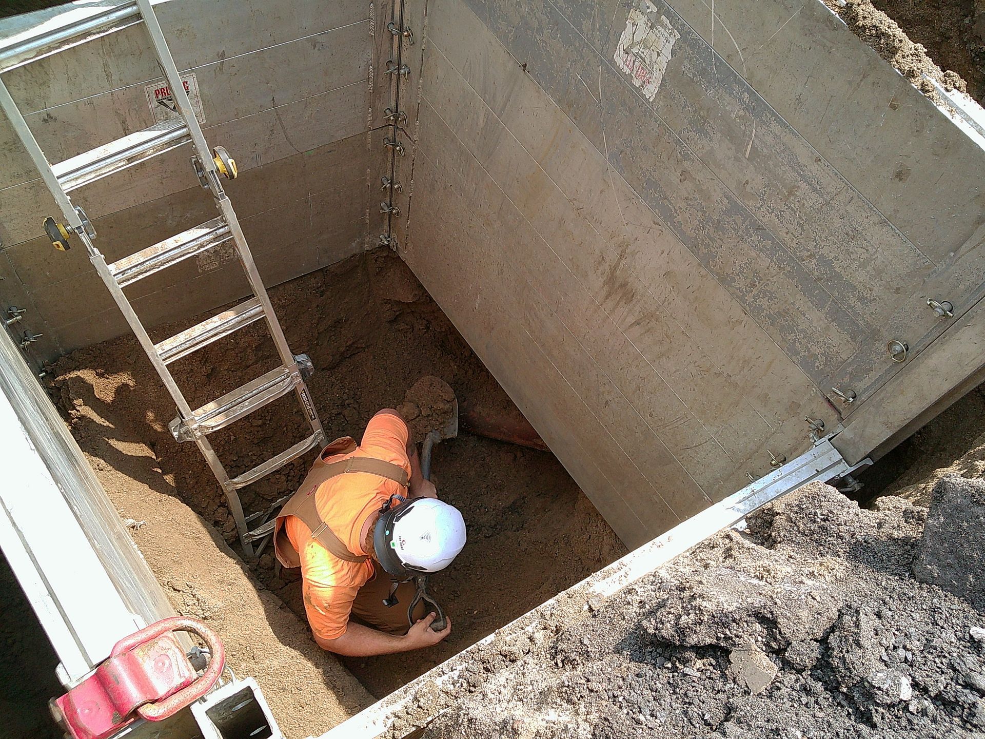 Worker in orange vest inside a rectangular trench, using a tool. Ladder is next to him.