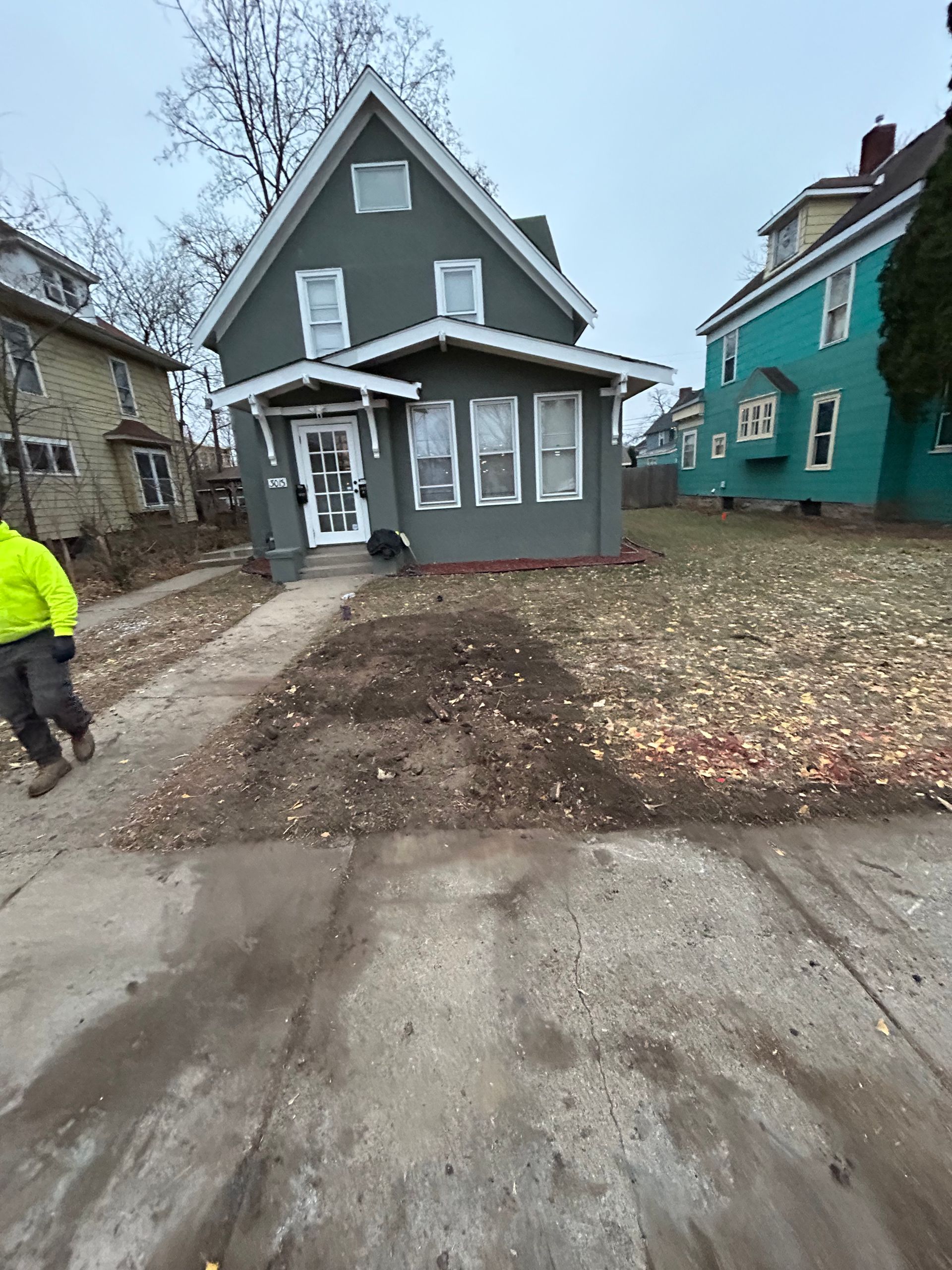 Green house with white trim; person walking left; yard bare and walkway in front.