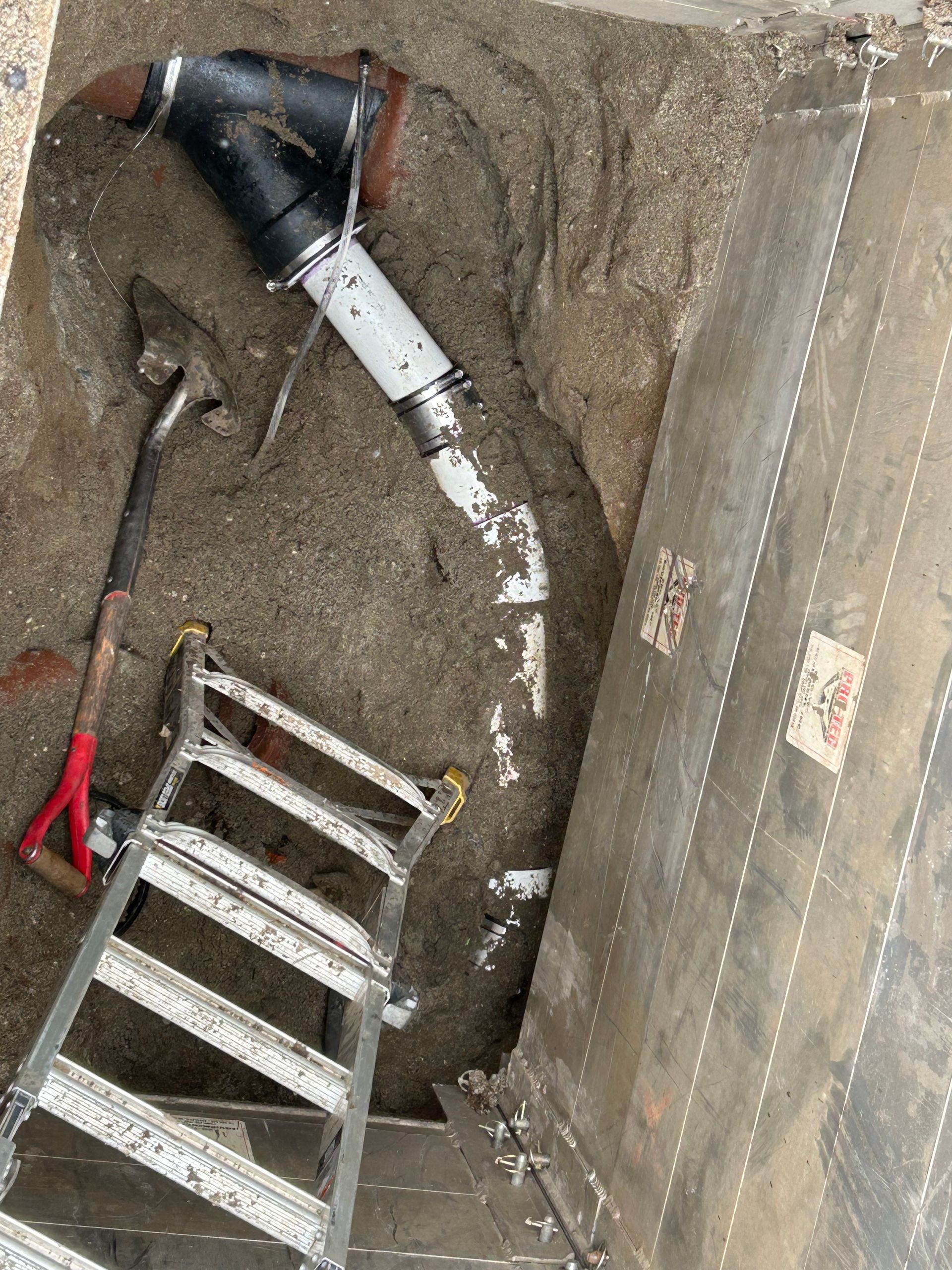 Plumbing repairs in a dirt pit. Black, white, and gray pipes connect in the ground near a ladder.