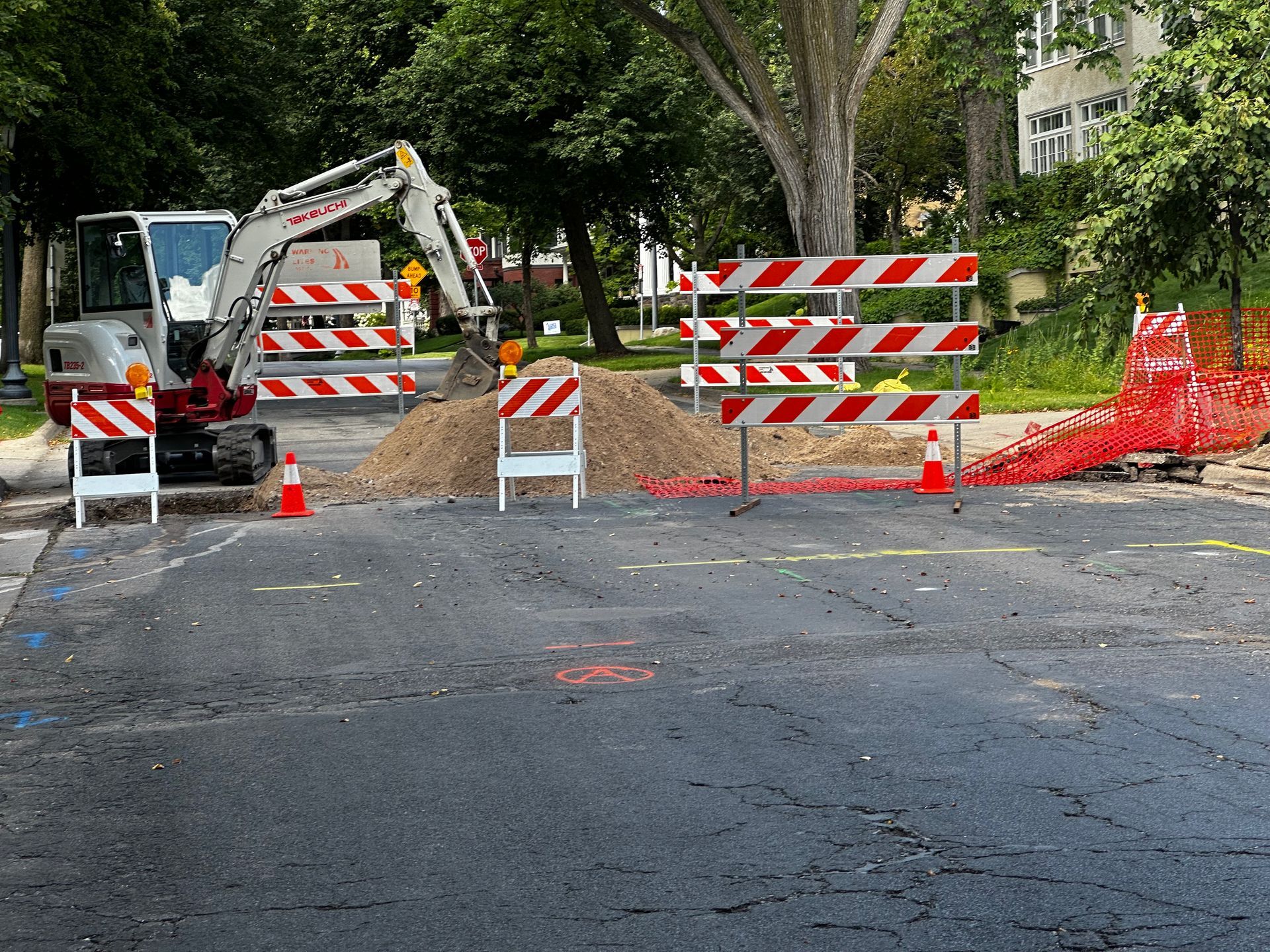 Road construction site: excavator, dirt pile, barricades, orange cones on asphalt.