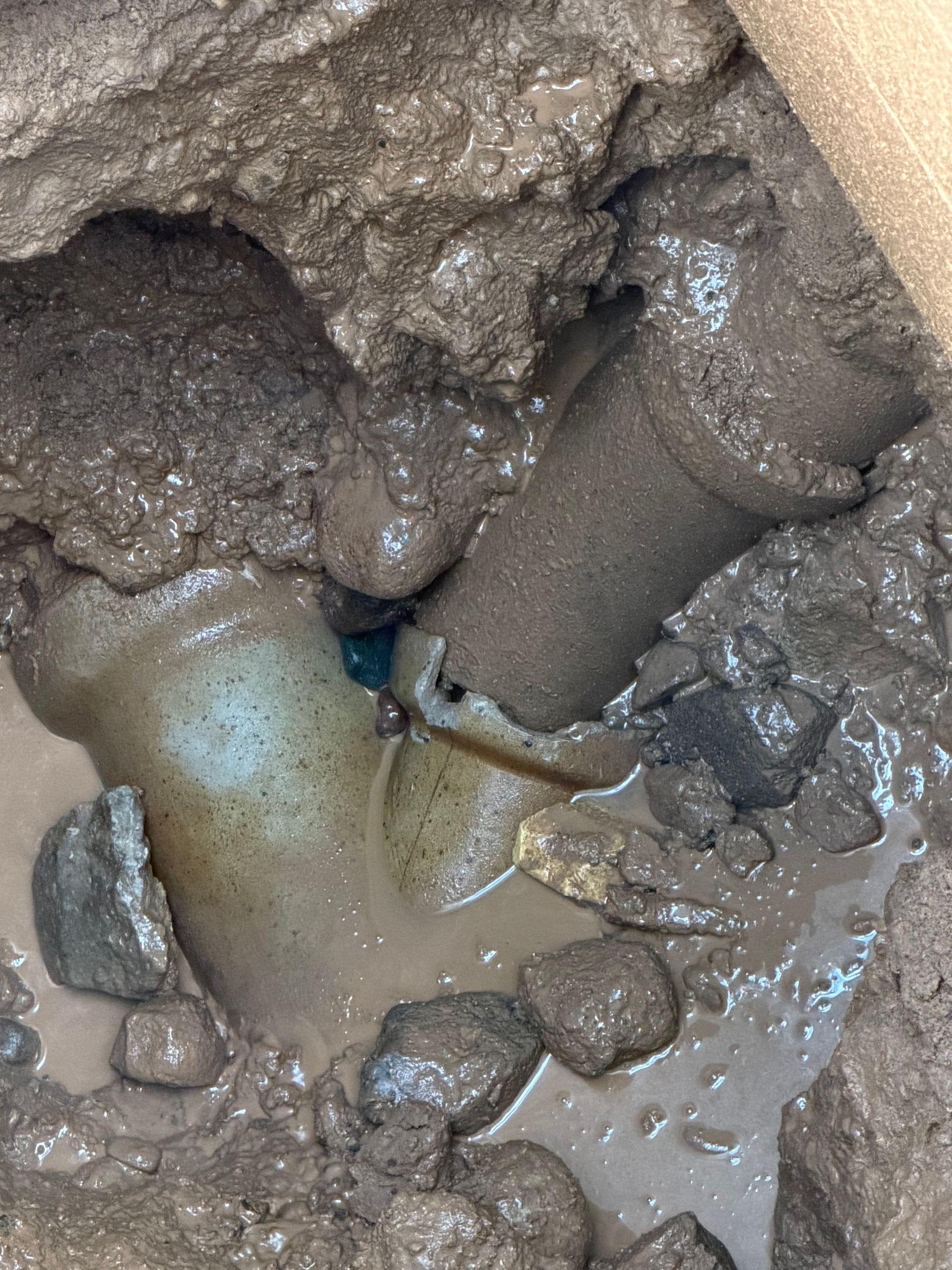 A broken pipe joint in mud. Blue-green discoloration where the joint has separated, surrounded by rocks and standing water.