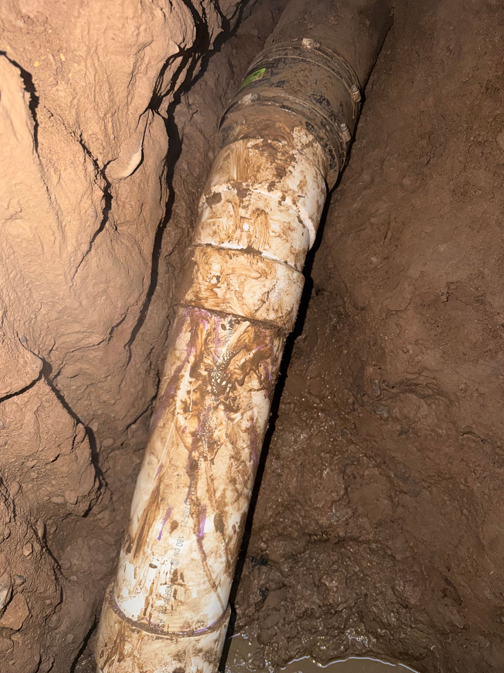 Damaged white PVC pipe section in a dirt trench. Brown dirt surrounds pipe with visible cracks and dirt stains.