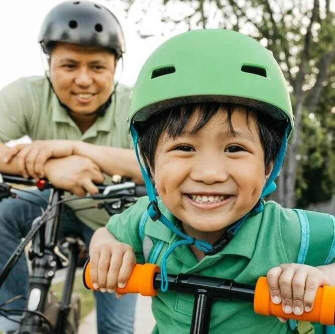 Smiling child in green helmet riding a scooter, with an adult cyclist behind outdoors