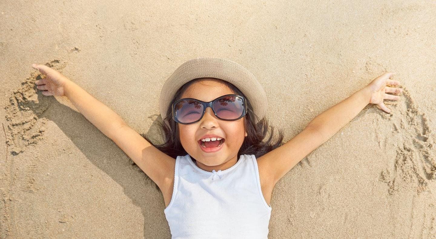Smiling child in sunglasses lying in sand with arms outstretched, making a sand angel