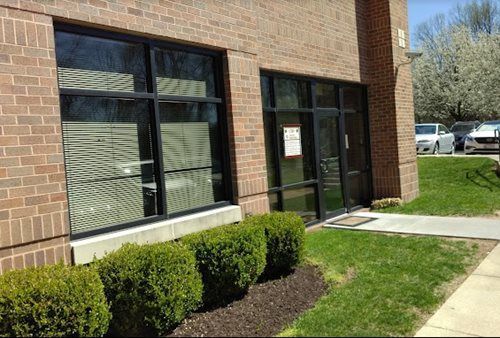 Brick building entrance with large windows, glass door, shrubs, and a sidewalk on a sunny day