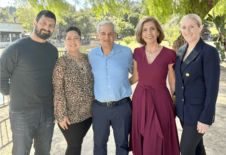 Five people standing outdoors, smiling in sunlight, with trees and houses in the background.