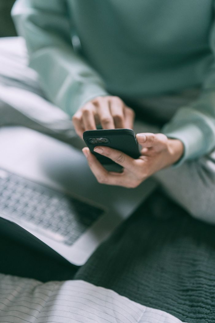 A person in a light green sweatshirt sits with a laptop, using their fingers to type on a smartphone.
