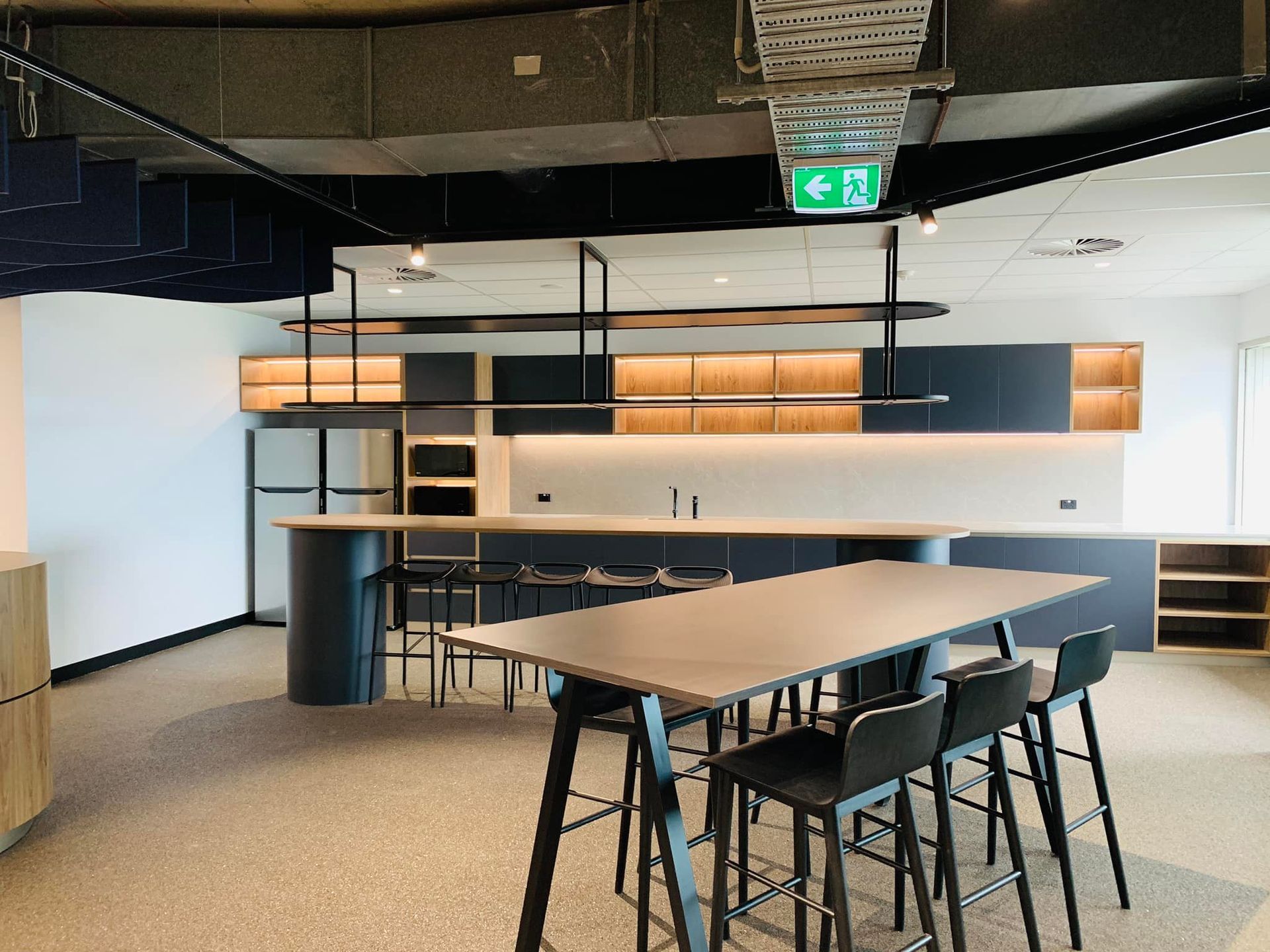 Modern office kitchenette with long table, stools, black counter, and open shelving under warm lights
