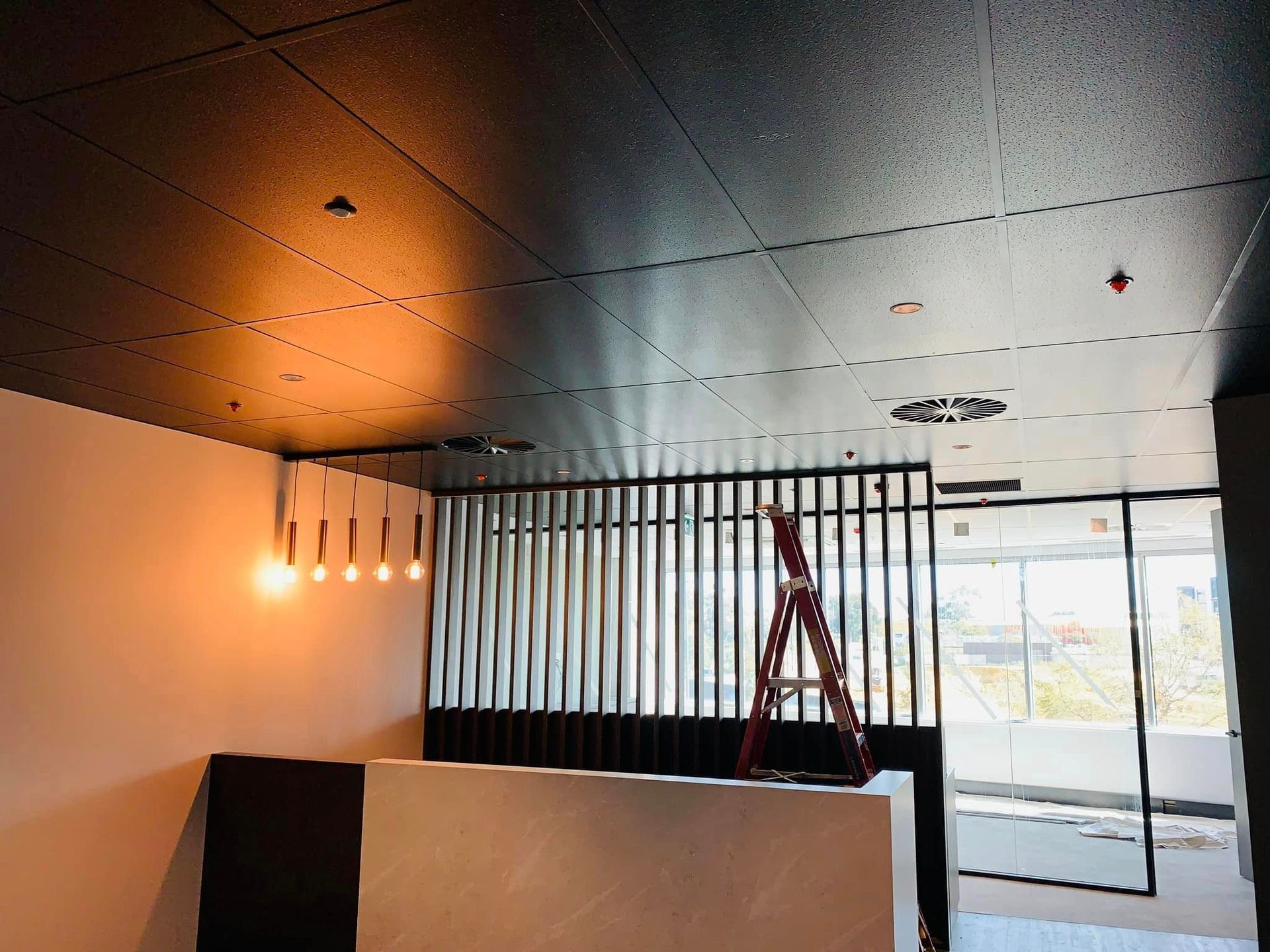Interior under construction with a ladder, exposed ceiling, and hanging lights near a window.