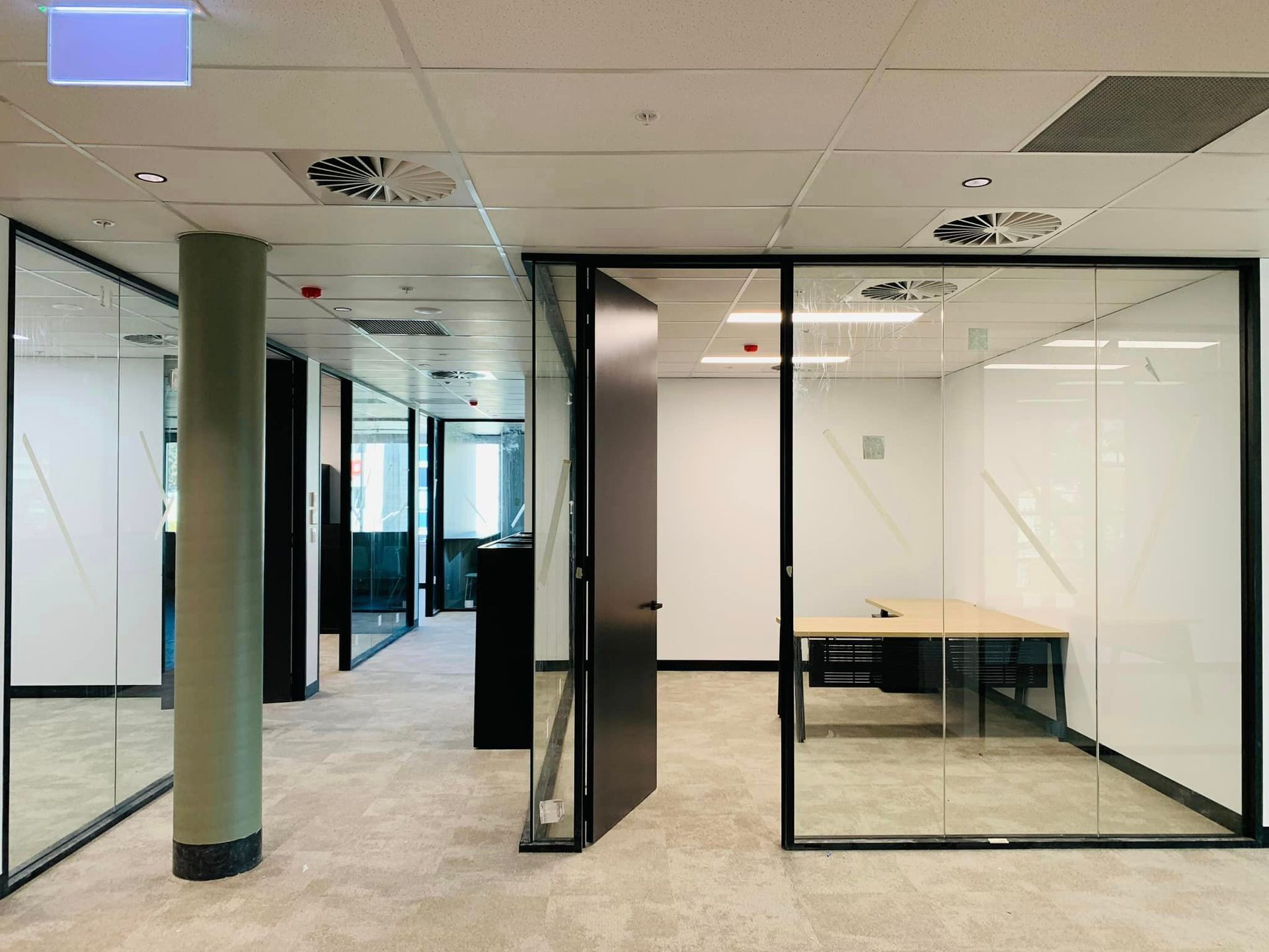 Empty office hallway with glass-walled rooms, desks, and green columns under a drop ceiling
