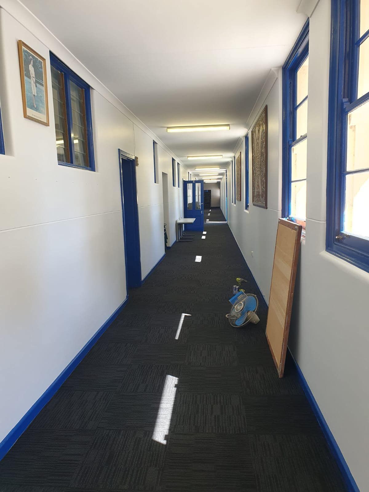 Empty school hallway with blue trim, windows on the right, and a backpack on the floor.