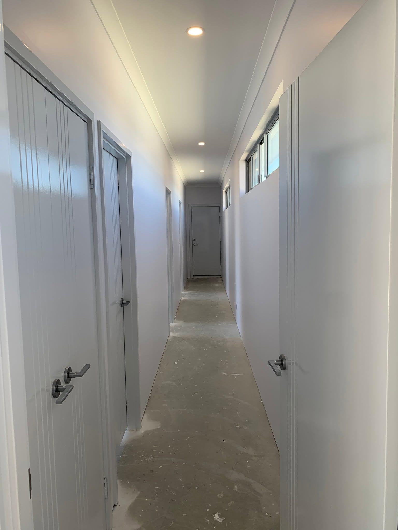 Narrow white hallway under renovation with doors, windows, and exposed concrete floor