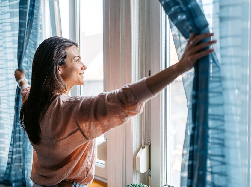 A woman is opening her curtains and looking out of a window.