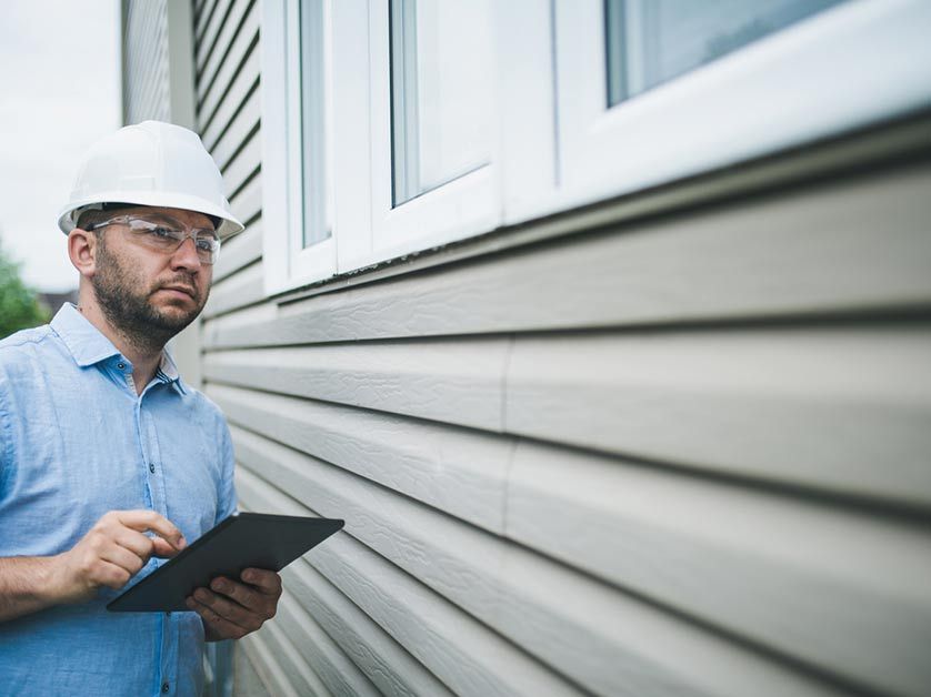 A man in a hard hat is looking at a tablet while standing next to a building.