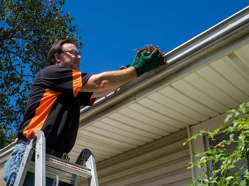 A man is standing on a ladder cleaning a gutter on a house.