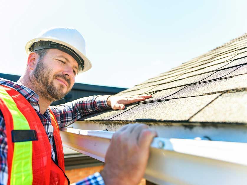 A man wearing a hard hat and safety vest is looking at a gutter on a roof.