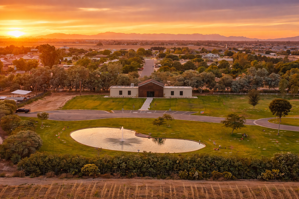 A single-story building sits by a small pond in a green park at sunset, with a neighborhood in the background.